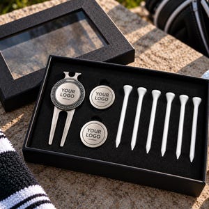 May include: A black gift box containing a silver metal golf divot tool, two circular metal ball markers, and six white golf tees. Each metal piece features the text "YOUR LOGO" in the centre. The box has a clear lid and black foam insert.