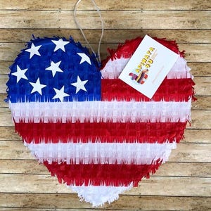 May include: Heart-shaped pi&ntilde;ata in the style of the American flag. The pi&ntilde;ata features red and white stripes with a blue section containing white stars. A white tag with the text "APINATA 50" is attached.