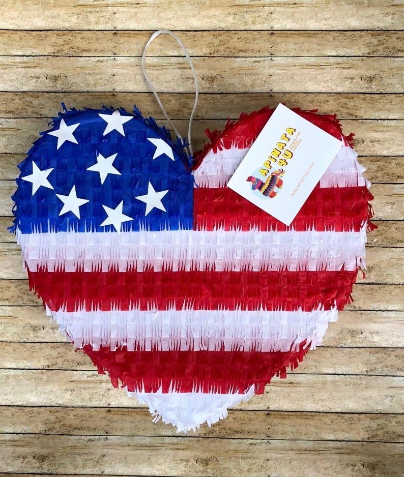 May include: Heart-shaped pi&ntilde;ata in the style of the American flag. The pi&ntilde;ata features red and white stripes with a blue section containing white stars. A white tag with the text "APINATA 50" is attached.