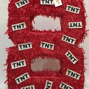 May include: A red number eight piñata covered in fringed paper, decorated with multiple white rectangular labels featuring the black text TNT. A small rectangular logo with a colorful piñata graphic and the text APINATA 4 U is attached.