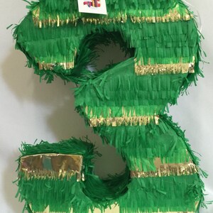 May include: A number three shaped piñata covered in green fringe paper with horizontal gold metallic strips. A white rectangular tag is attached to the top, featuring the text APINATA 4U and a small graphic of a colorful piñata on a white background.