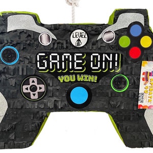 May include: A black and silver piñata shaped like a video game controller, with the words "GAME ON! YOU WIN!" in white and green. It features colorful buttons and a rope for hanging. The piñata is designed for a party or celebration.