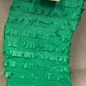 May include: A green cactus-shaped pi&ntilde;ata with fringed paper detailing. A colorful label with the text "APi&ntilde;ata" and a QR code is attached. Yellow paper flowers and a white string are at the top.