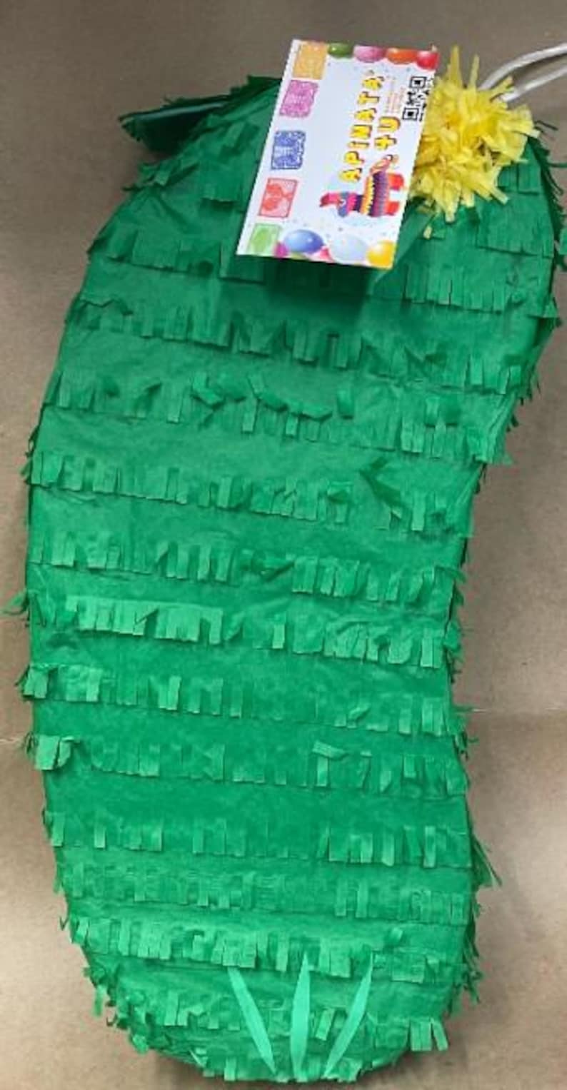 May include: A green cactus-shaped pi&ntilde;ata with fringed paper detailing. A colorful label with the text "APi&ntilde;ata" and a QR code is attached. Yellow paper flowers and a white string are at the top.