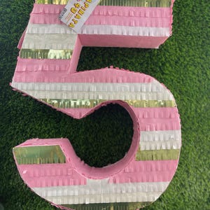 May include: A large, pink number five piñata with alternating white and gold stripes. The piñata is decorated with fringed paper and has a tag that reads "Piñata 4 U". The piñata is on a green surface.