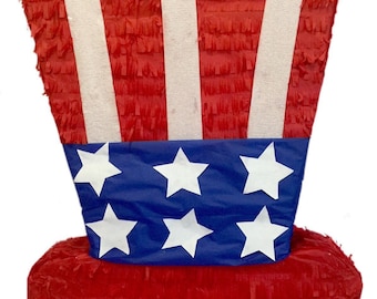 May include: A red, white, and blue Uncle Sam hat piñata. The hat features red and white stripes on the top, a blue band with white stars, and a red brim. The piñata is made of paper and is designed for a festive occasion.