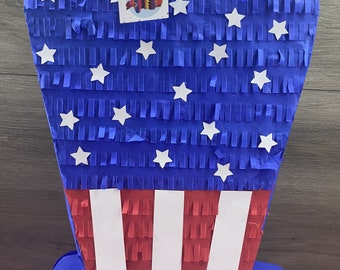 May include: A blue and red pinata shaped like a top hat with white stars. The pinata has a red, white, and blue striped band around the middle.