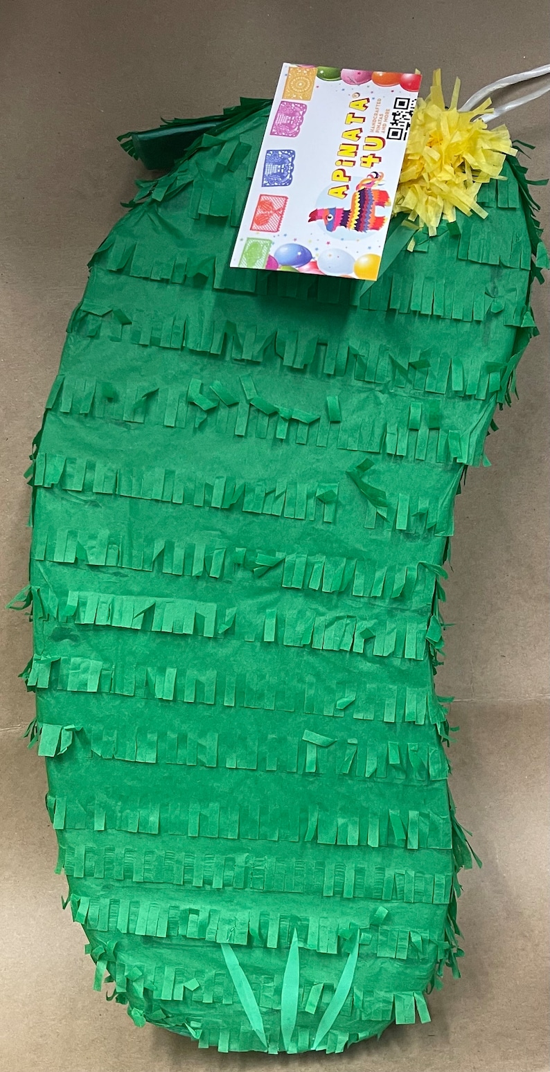 May include: A green cactus-shaped pi&ntilde;ata with fringed edges, a yellow flower, and a tag that reads "Apinata 4 U". The pi&ntilde;ata is made of paper and is designed for festive celebrations.