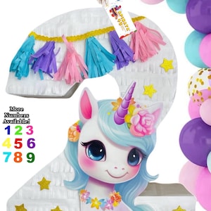 May include: A number 2 piñata with a unicorn design. The piñata is white with gold star accents and colorful fringe. A unicorn with blue hair, a flower crown, and a floral necklace is featured on the front. Balloons in various colors surround the piñata.