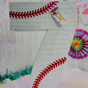 May include: A white number seven piñata with red baseball stitching detail. The piñata has a tag that reads "APINATA 4U". The background includes party decorations.