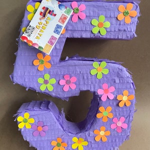 May include: A large, purple number five piñata decorated with colorful paper flowers. The piñata has a fringed texture and a small tag with text and a QR code. The flowers are in shades of pink, orange, yellow, and green.