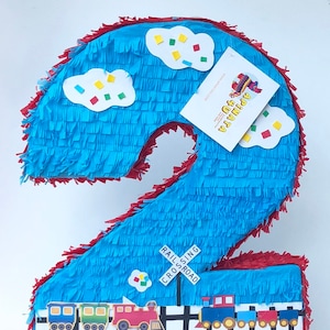 May include: A large, blue number two piñata with red trim, decorated with white clouds, colorful confetti, and a train scene. The piñata has a railroad crossing sign and a business card attached.
