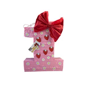 May include: Pink number one shaped pinata with a red bow and strawberry and daisy decorations. The pinata has a tag that says "Oh, the places you'll go!"