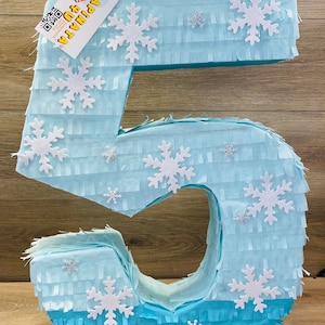 May include: A light blue number 5 piñata decorated with white glitter snowflakes. The piñata is made of fringed paper and has a tag that reads "A Pinata 4 U". This festive decoration is perfect for a birthday party.