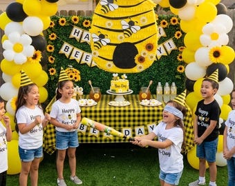 May include: A bee-themed party setup featuring a large yellow and black beehive piñata, a balloon arch with sunflowers, a table with a yellow and black checkered cloth, a cake, and children wearing party hats and shirts that say Happy Bee-Day.