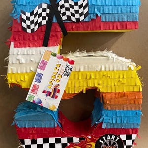 May include: A colorful number 5 piñata with a racing theme. The piñata is decorated with blue, red, white, yellow, and orange fringed paper. It features checkered flags, a racing flame graphic, and a sticker that says "Start Your Engines!"