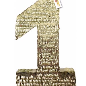 May include: A large, gold-colored number one piñata, constructed with layered, fringed material. The piñata has a rectangular base and a tag attached to the top. The overall design is festive and celebratory.