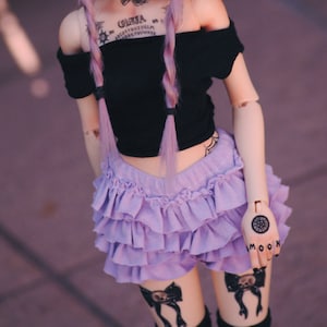 Könnte beinhalten: Eine Puppe mit lila Haaren, die ein schwarzes, schulterfreies Oberteil, einen violetten, gerüschten Rock und einen schwarzen Hut trägt. Die Puppe hat Tätowierungen auf ihrer Brust und ihren Armen, darunter ein Ouija-Brett und einen Halbmond.