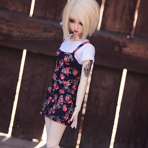 May include: A doll with blonde hair wearing a floral dress, a white t-shirt, and black combat boots. The doll has a skull tattoo on its arm and a tattoo on its face that says "Wild Child".