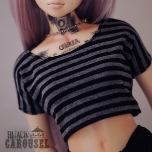 Puede incluir: Una muñeca con cabello morado que lleva un top corto a rayas negras y grises. La muñeca tiene tatuajes en el cuello y el pecho, incluyendo la palabra "Ouija" y un diseño de una tabla Ouija.