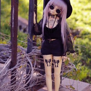 May include: A doll with long purple hair wearing a black hat, sunglasses, a black top, and black shorts. The doll has tattoos on its legs and arms. The doll is standing in front of a metal fence.