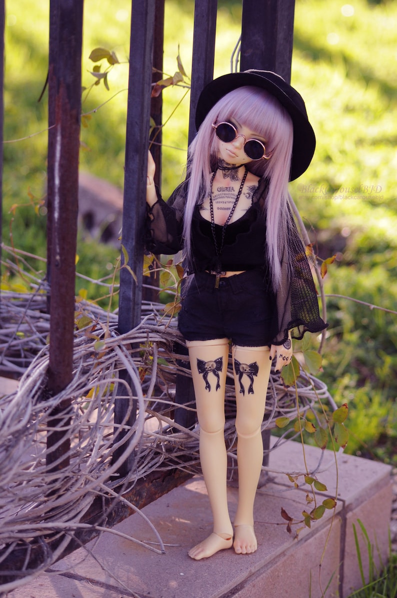 May include: A doll with long purple hair wearing a black hat, sunglasses, a black top, and black shorts. The doll has tattoos on its legs and arms. The doll is standing in front of a metal fence.