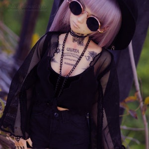 May include: A doll wearing a black mesh top with a black velvet choker and a black hat. The doll has a purple wig and is wearing black shorts. The doll has several tattoos, including a Ouija board, a crescent moon, and a sun.
