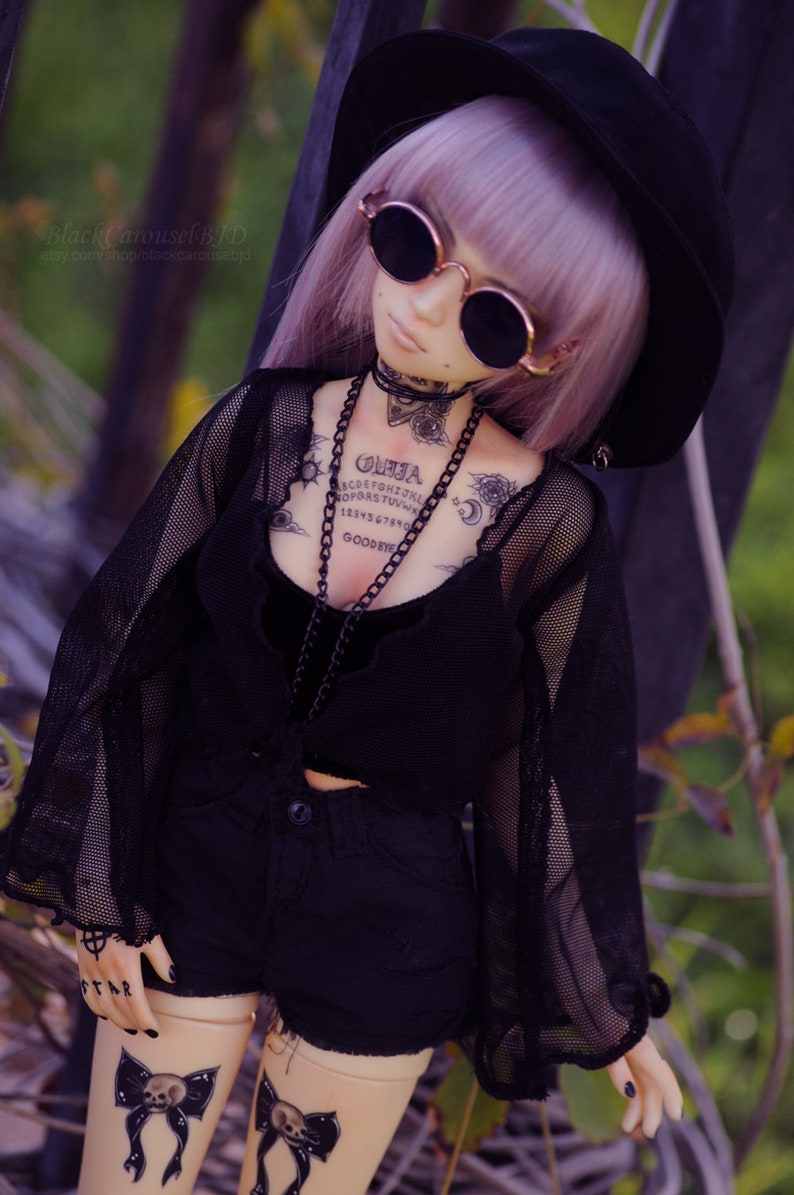 May include: A doll wearing a black mesh top with a black velvet choker and a black hat. The doll has a purple wig and is wearing black shorts. The doll has several tattoos, including a Ouija board, a crescent moon, and a sun.