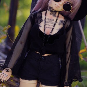 May include: A doll with purple hair wearing a black mesh top, black shorts, and a black hat. The doll has several tattoos, including a Ouija board, a skull, and a crescent moon. The doll is wearing round black sunglasses.
