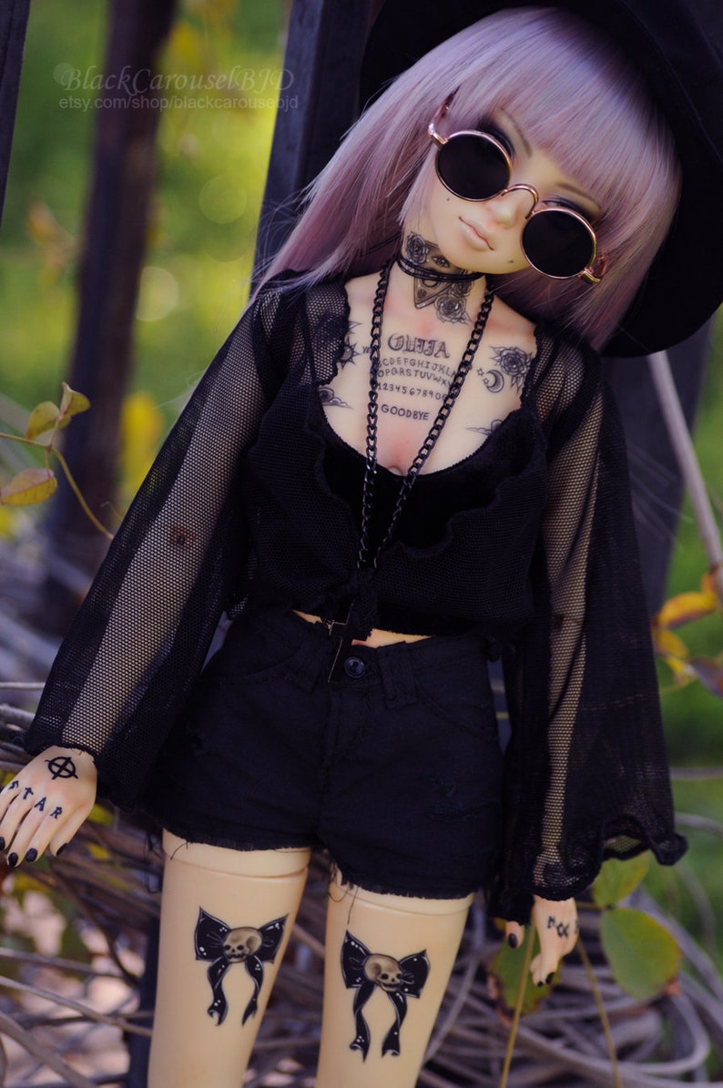 May include: A doll with purple hair wearing a black mesh top, black shorts, and a black hat. The doll has several tattoos, including a Ouija board, a skull, and a crescent moon. The doll is wearing round black sunglasses.
