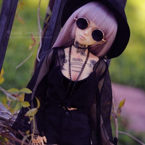 May include: A doll with purple hair wearing a black hat, sunglasses, a black mesh top, black shorts, and black and white tattoos. The doll has a Ouija board tattoo on its chest.
