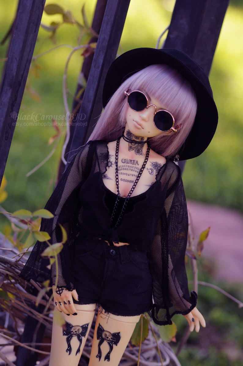 May include: A doll with purple hair wearing a black hat, sunglasses, a black mesh top, black shorts, and black and white tattoos. The doll has a Ouija board tattoo on its chest.