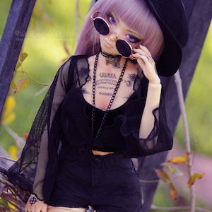 May include: A doll with purple hair wearing a black hat, sunglasses, and a black mesh top with a black chain necklace. The doll has tattoos on its chest and thighs, including a Ouija board and a skull and bow.