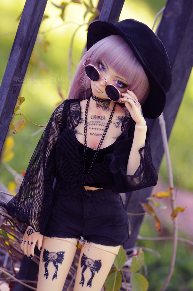 May include: A doll with purple hair wearing a black hat, sunglasses, and a black mesh top with a black chain necklace. The doll has tattoos on its chest and thighs, including a Ouija board and a skull and bow.