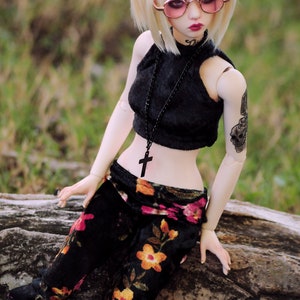 May include: A doll with blonde hair wearing a black halter top with a black beaded necklace and a cross pendant. The doll is also wearing black pants with a floral pattern and pink-tinted round sunglasses. The doll has a skull tattoo on its arm.