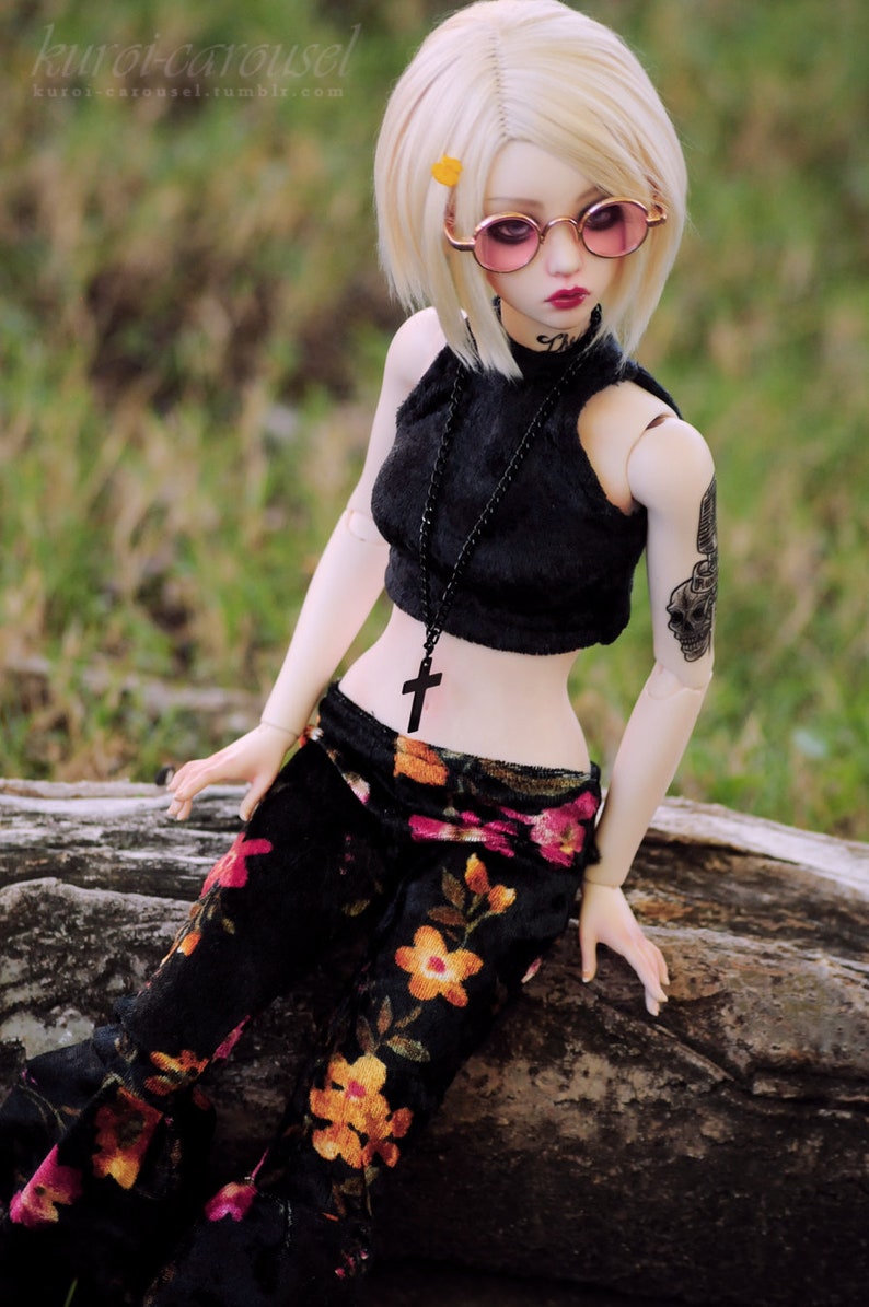 May include: A doll with blonde hair wearing a black halter top with a black beaded necklace and a cross pendant. The doll is also wearing black pants with a floral pattern and pink-tinted round sunglasses. The doll has a skull tattoo on its arm.