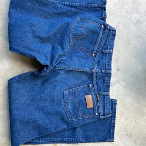 May include: A pair of blue denim jeans with two back pockets and a brown leather patch with the word "Wrangler" on the right back pocket.