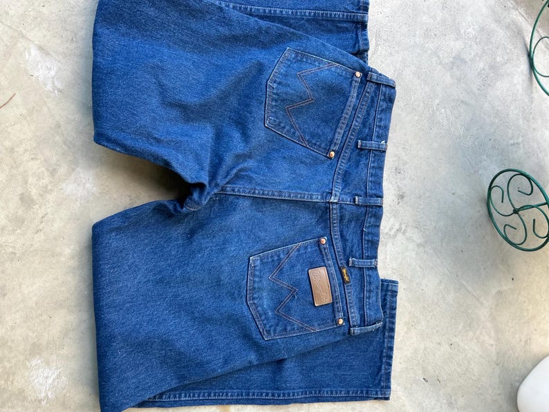 May include: A pair of blue denim jeans with two back pockets and a brown leather patch with the word "Wrangler" on the right back pocket.
