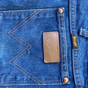 May include: Close-up of a blue denim back pocket with a brown leather Wrangler label. The pocket has distinctive W-shaped stitching.