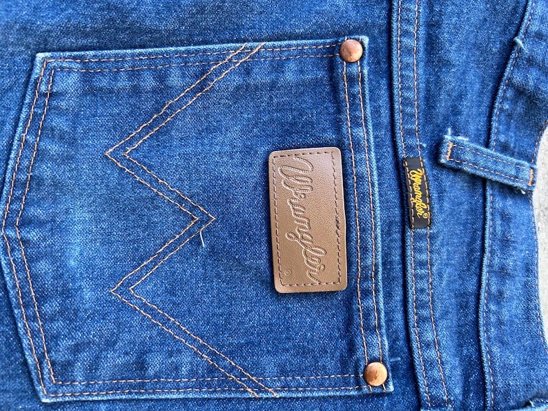 May include: Close-up of a blue denim back pocket with a brown leather Wrangler label. The pocket has distinctive W-shaped stitching.