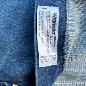 May include: A blue denim Wrangler jeans label with white text. The label states "Wrangler 13MWZPW 38 X 30" and includes care instructions, size, and manufacturing information.