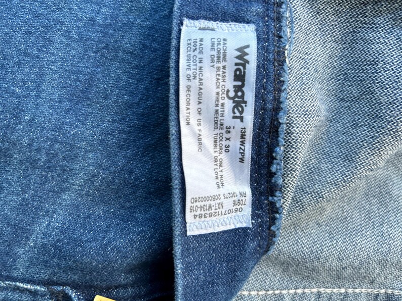 May include: A blue denim Wrangler jeans label with white text. The label states "Wrangler 13MWZPW 38 X 30" and includes care instructions, size, and manufacturing information.