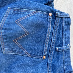 May include: Close-up of the back pocket of a pair of blue denim jeans. The pocket has a distinctive orange stitching detail in the shape of a 'W'.