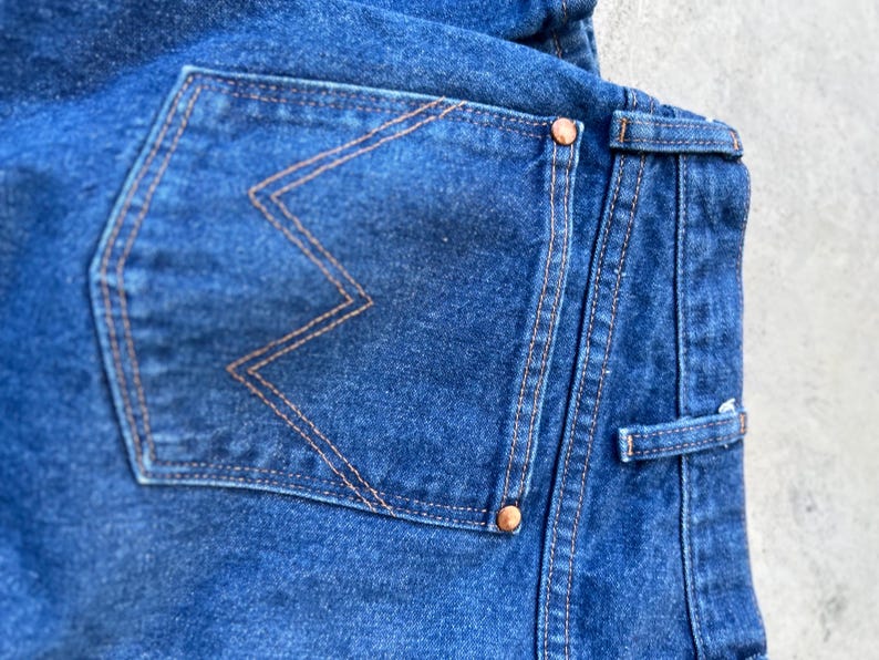 May include: Close-up of the back pocket of a pair of blue denim jeans. The pocket has a distinctive orange stitching detail in the shape of a 'W'.
