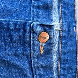 May include: Close-up of a blue denim jean with a copper button. The button has a small, intricate design.