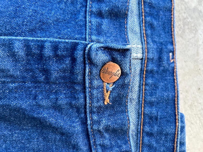 May include: Close-up of a blue denim jean with a copper button. The button has a small, intricate design.