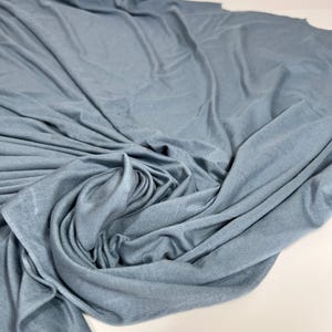 May include: A large piece of blue fabric, possibly a textile or cloth, is draped and folded, creating soft, flowing lines. The fabric has a slightly textured appearance and a muted, dusty blue color. The folds and shadows add depth and dimension to the material.