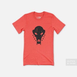 May include: Coral-colored t-shirt featuring a distressed black graphic of a stylized skull with star accents. The shirt has short sleeves and a crew neck. The text "ALPHA K-NINE" is in the lower right corner.