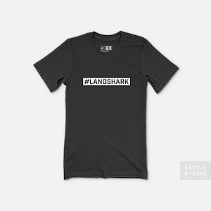 May include: A black t-shirt with a white rectangle on the front that says "#LANDSHARK".