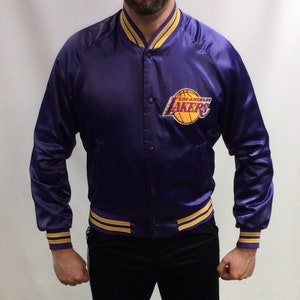 May include: A purple satin bomber jacket with yellow and gold stripes on the cuffs and waistband. The jacket has a large Los Angeles Lakers logo on the left chest.